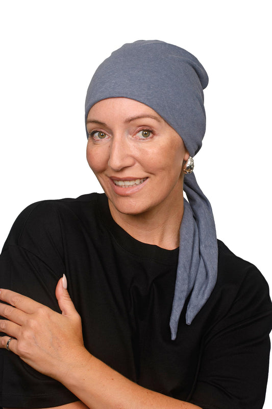 A woman wearing a slate blue fabric cancer head covering or scarf with long hanging tails, styled over her shoulders, set against a plain white background.