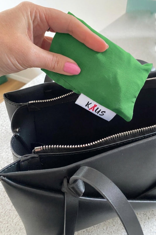 A hand with pink nail polish is placing a green fabric pouch into a black zippered handbag. The inside of the bag features a white fabric label with the brand name 'KAUS' in red and black text.