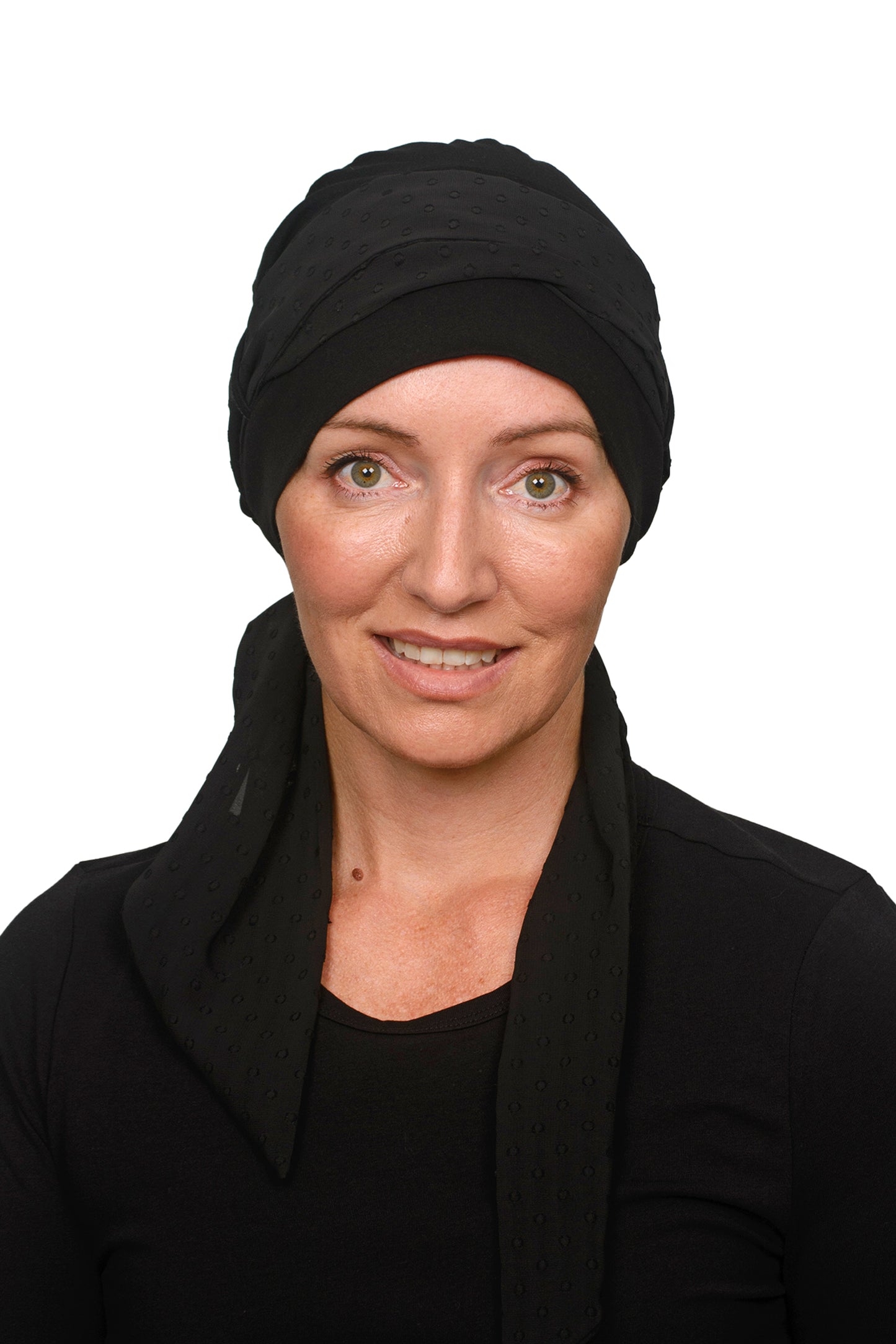 A smiling woman with green eyes wearing a black headscarf for cancer and black long-sleeve top, resting her chin on her hand against a solid white background.