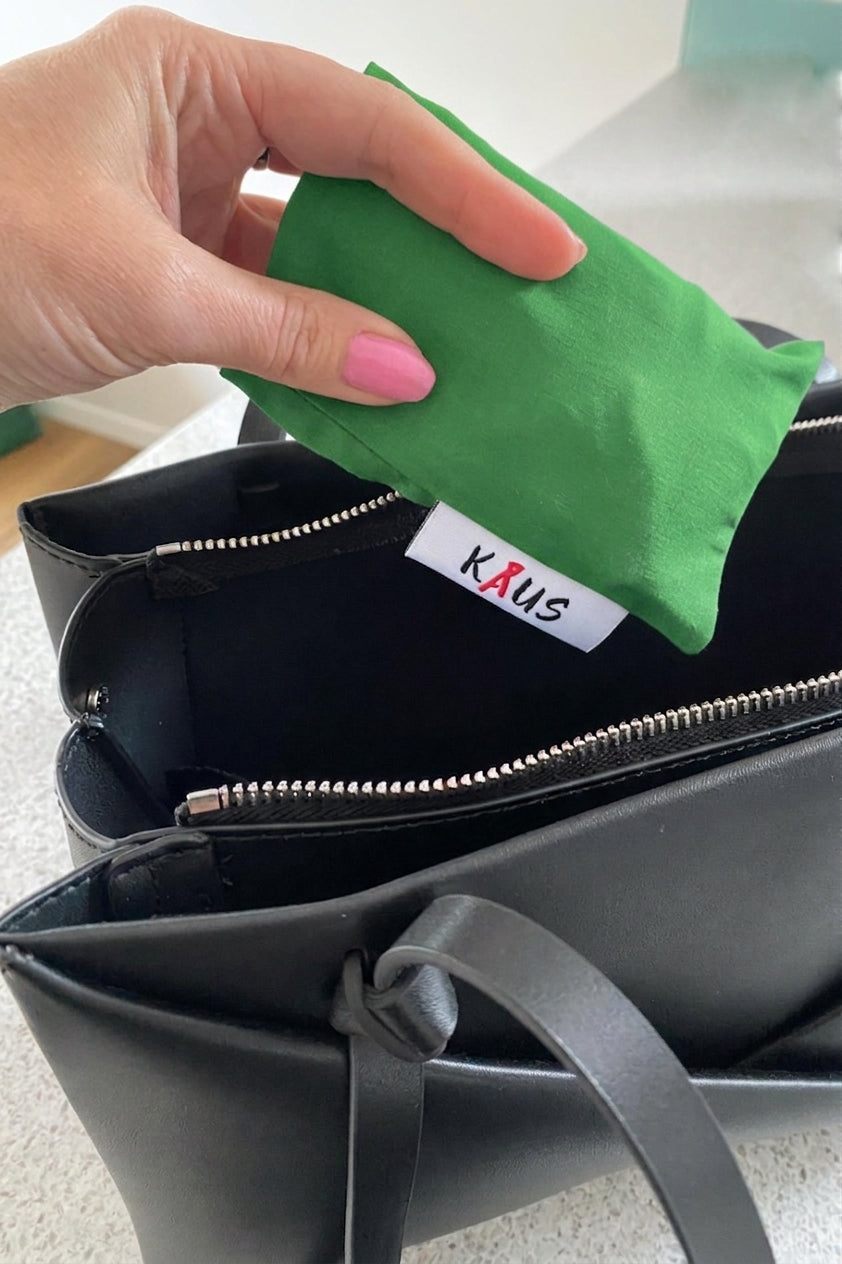 A hand with pink nail polish is placing a green fabric pouch into a black zippered handbag. The inside of the bag features a white fabric label with the brand name 'KAUS' in red and black text.
