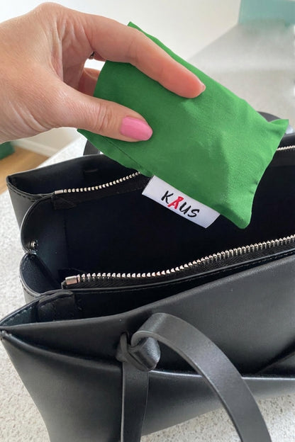 A hand with pink nail polish is placing a green fabric pouch into a black zippered handbag. The inside of the bag features a white fabric label with the brand name 'KAUS' in red and black text.