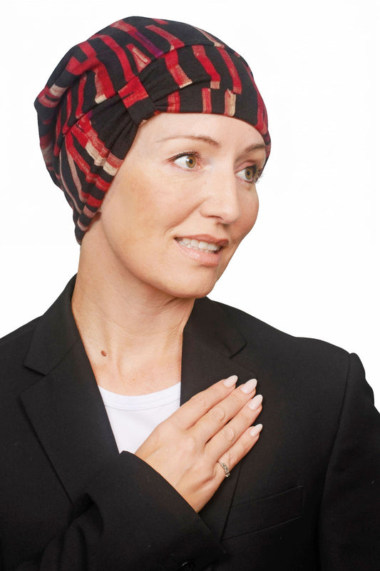 A woman wearing a black and red patterned headscarf, a black blazer, and a white undershirt, with her hand over her chest, set against a plain white background.