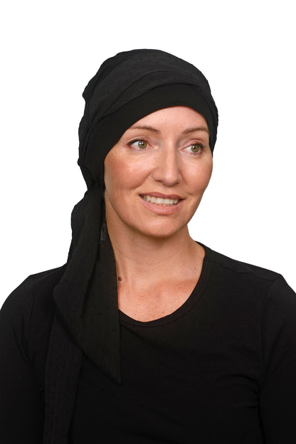 A smiling woman with green eyes wearing a black headscarf for cancer and black long-sleeve top, resting her chin on her hand against a solid white background.
