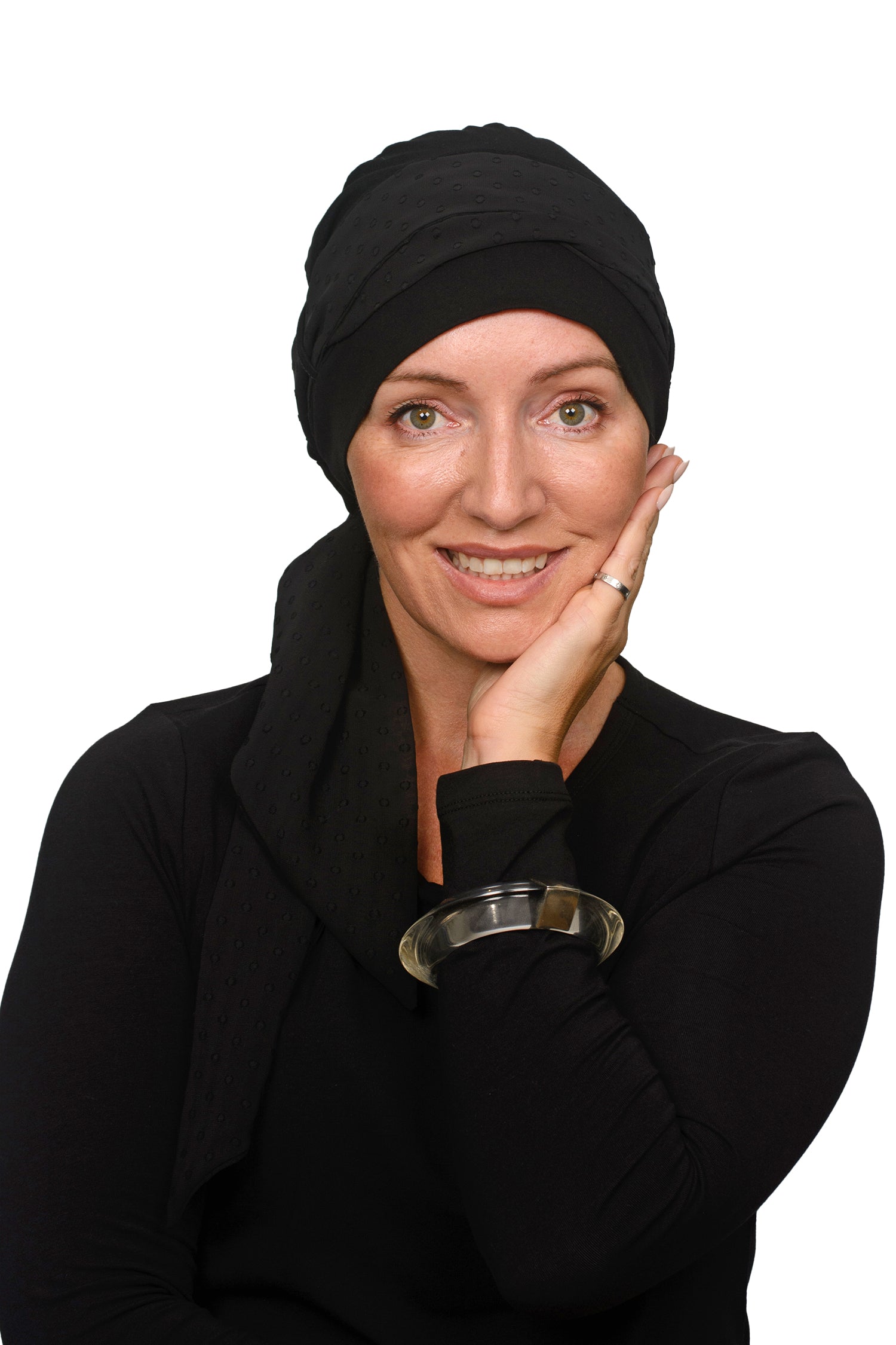 A smiling woman with green eyes wearing a black headscarf for cancer and black long-sleeve top, resting her chin on her hand against a solid white background.